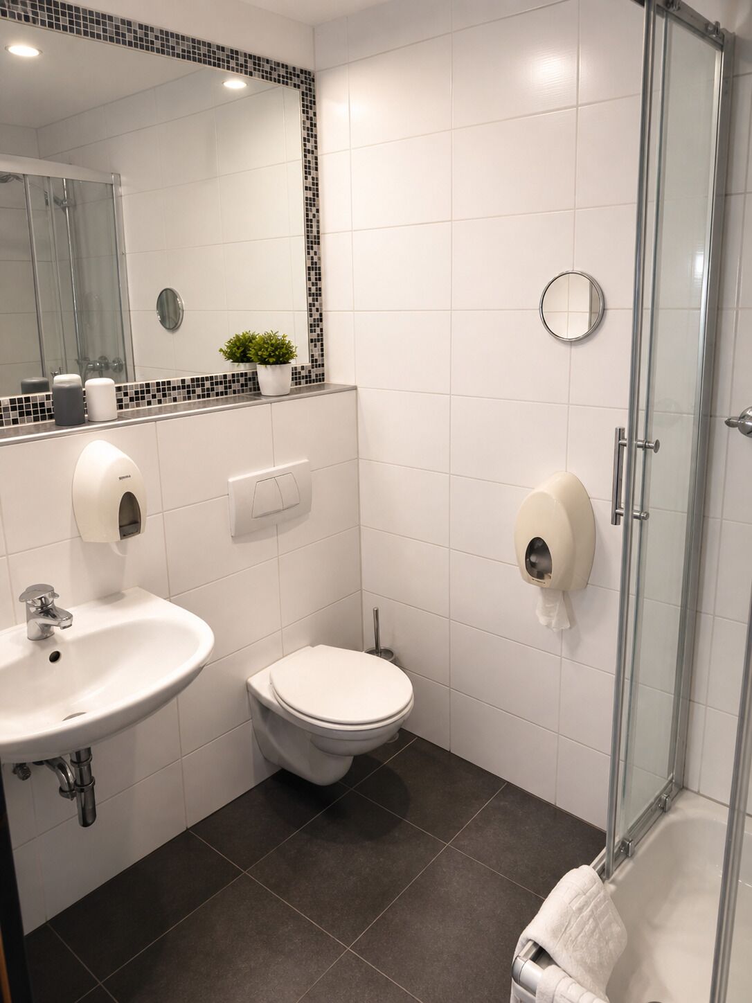 Un baño moderno con cabina de ducha de vidrio, un lavabo blanco, un inodoro y un espejo con detalles de mosaico.