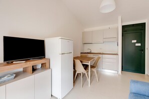 Ein kompakter Wohnraum mit Fernseher auf einem Holzgestell, einer kleinen Küchenzeile inklusive Kühlschrank sowie einem Esstisch mit Stühlen.