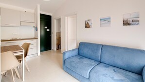 A modern living room with a blue sofa, a dining table with chairs, a kitchen area, and a door leading to another room. - Apartment with sea view terrace and air conditioning (Castelsardo)