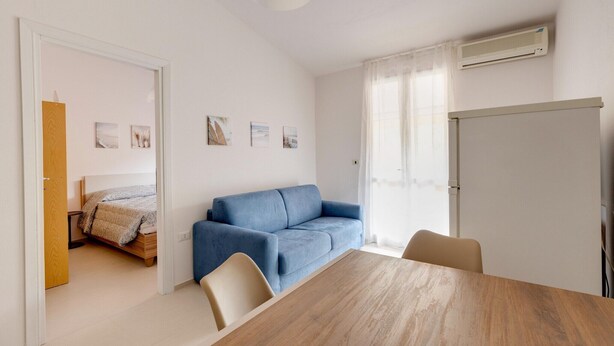 A room with a wooden dining table, a blue sofa, a bed with a wooden headboard, and a white air conditioning unit on the wall. - Apartment with sea view terrace and air conditioning (Castelsardo)