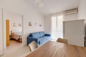 A room with a wooden dining table, a blue sofa, a bed with a wooden headboard, and a white air conditioning unit on the wall. - Apartment with sea view terrace and air conditioning (Castelsardo)