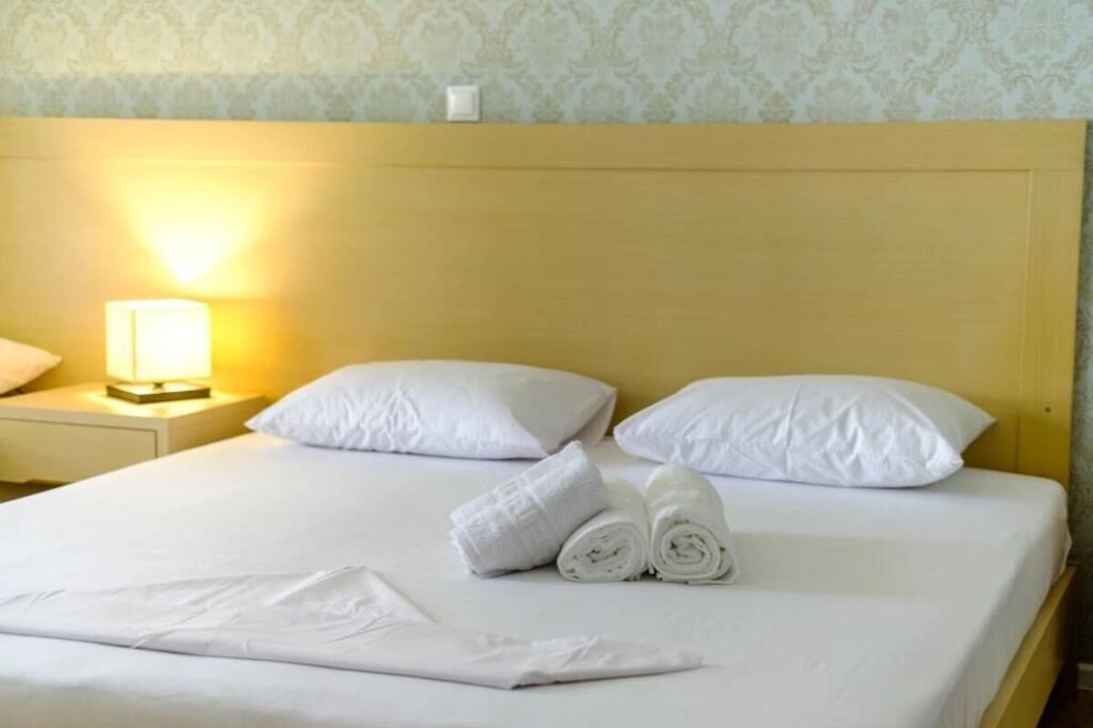 A hotel room with a neatly made bed, white pillows, rolled towels, and a bedside lamp.