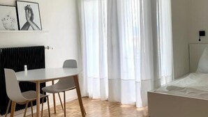 Un dormitorio moderno con una cama, una mesa de comedor con sillas, un radiador y una ventana con cortinas translúcidas.