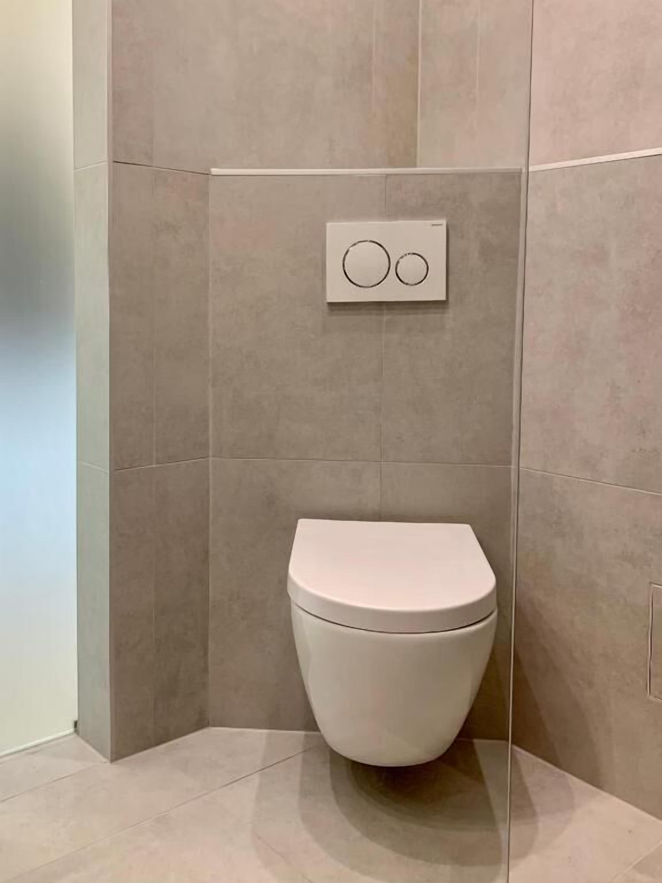 Une salle de bain moderne avec des toilettes blanches, des carreaux beiges et un panneau de commande de chasse d’eau fixé au mur.