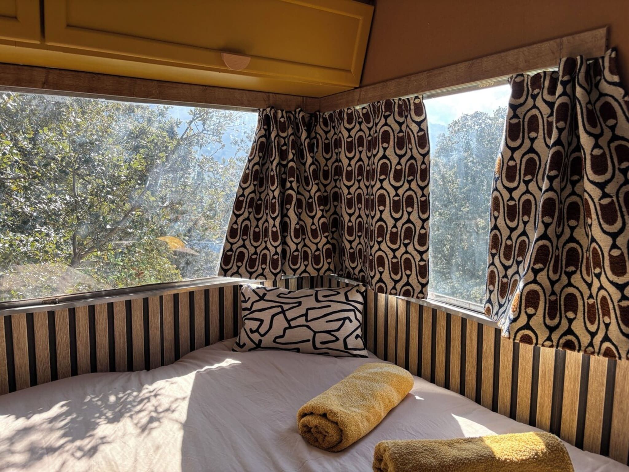 A bed with a patterned pillow, a rolled towel, and a window with patterned curtains.