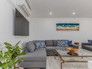 A modern living room with a grey sectional sofa, a glass coffee table, and a painting of a beach scene. - Coastal Luxe Rooftop Escape - Paradiso 345 (Tweed)