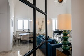 Ein modernes Wohnzimmer mit Glastrennwand, einem blauen Sofa, einem Essbereich mit Tisch und Stühlen sowie einem Klavier.
