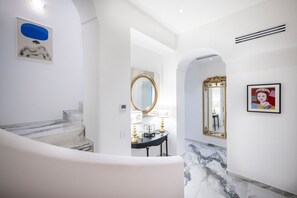 Ein modernes Interieur mit einer Marmortreppe, einem runden Spiegel, einem Konsolentisch mit Lampen und einem gerahmten Kunstwerk an der Wand.
