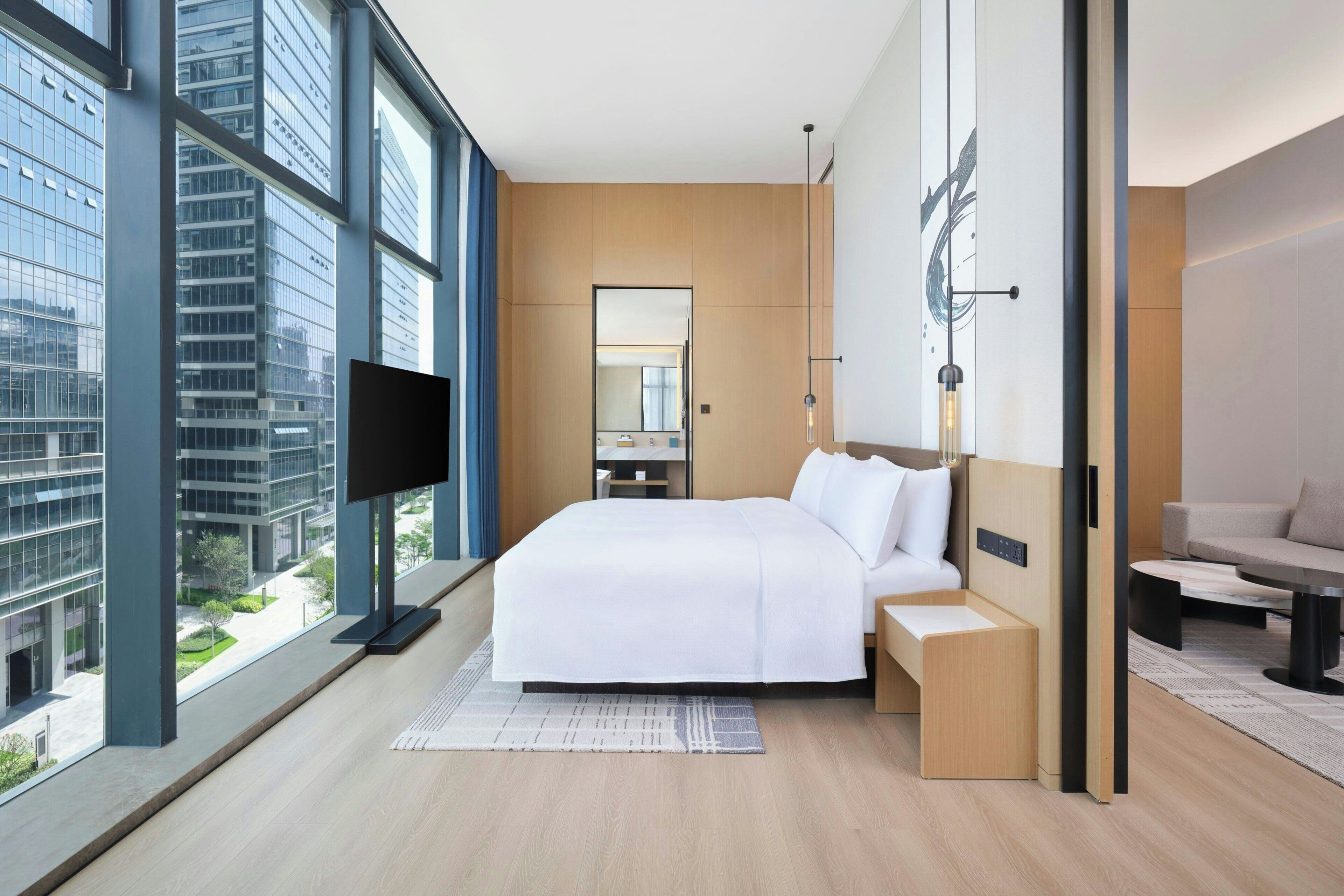 A modern hotel room with a large bed, a TV, a desk, and a view of the cityscape through large windows.