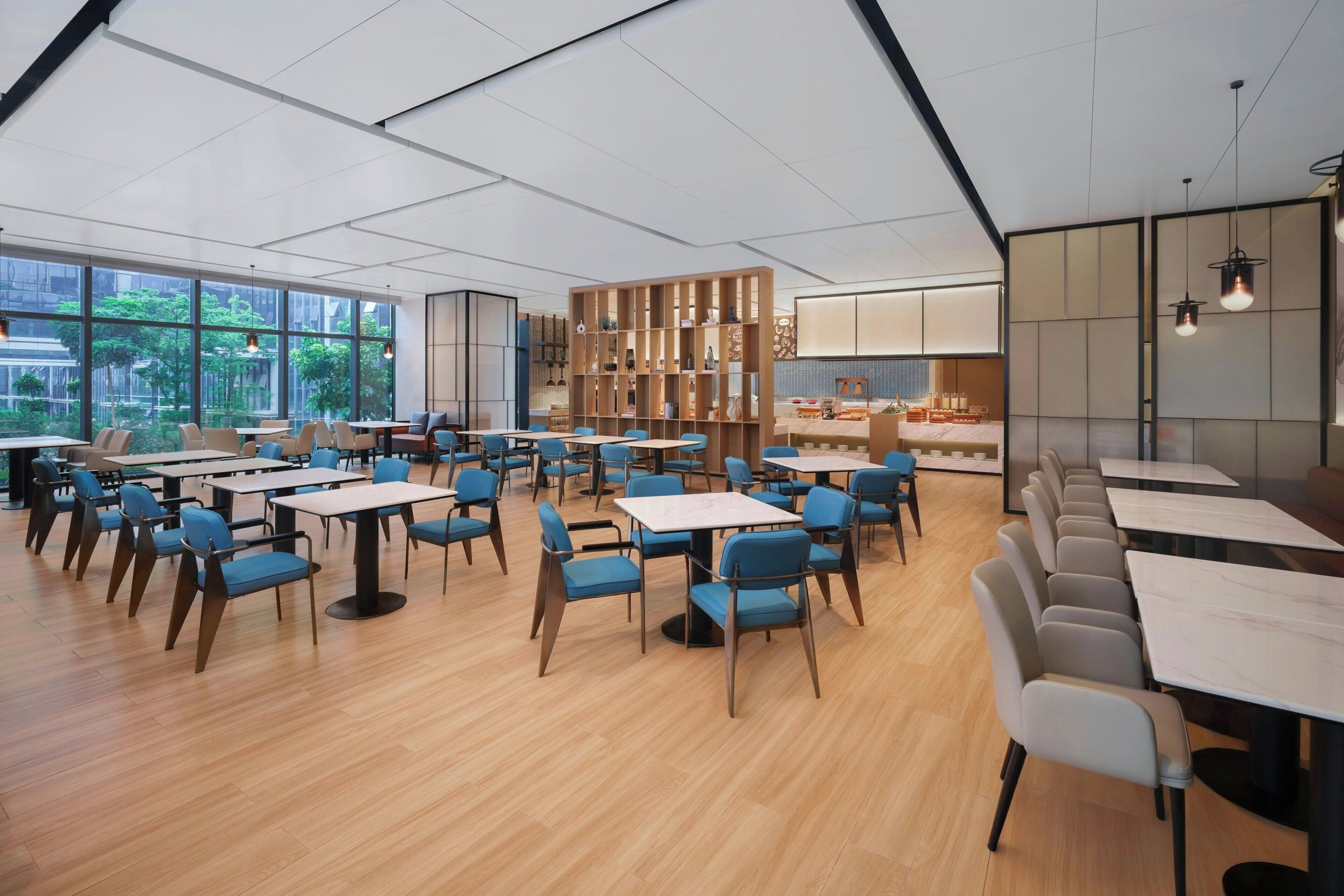 Een moderne eethoek met tafels en stoelen, grote ramen en uitzicht op groen.