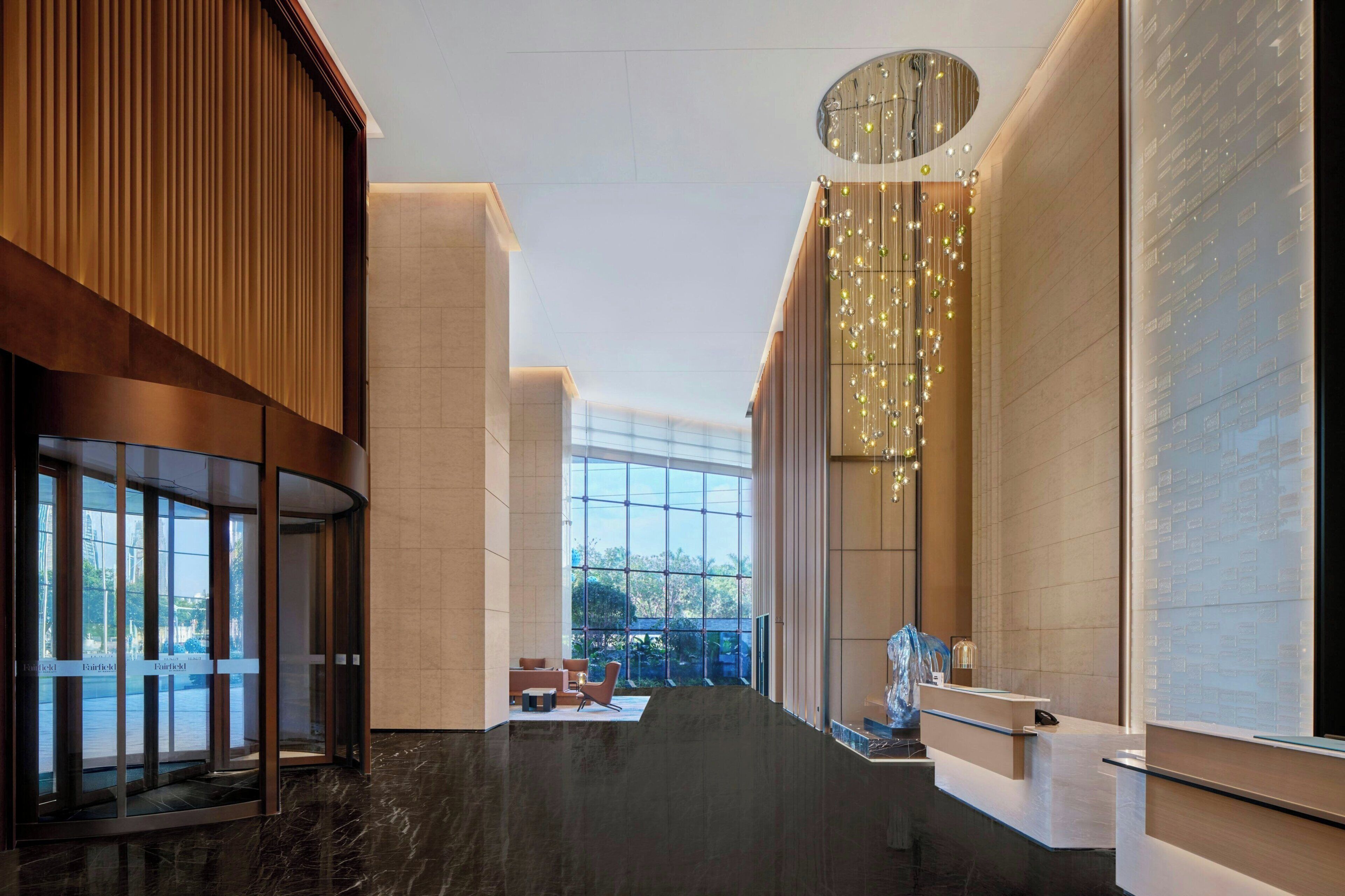 A modern hotel lobby with a glass elevator, seating area, and a decorative light fixture.