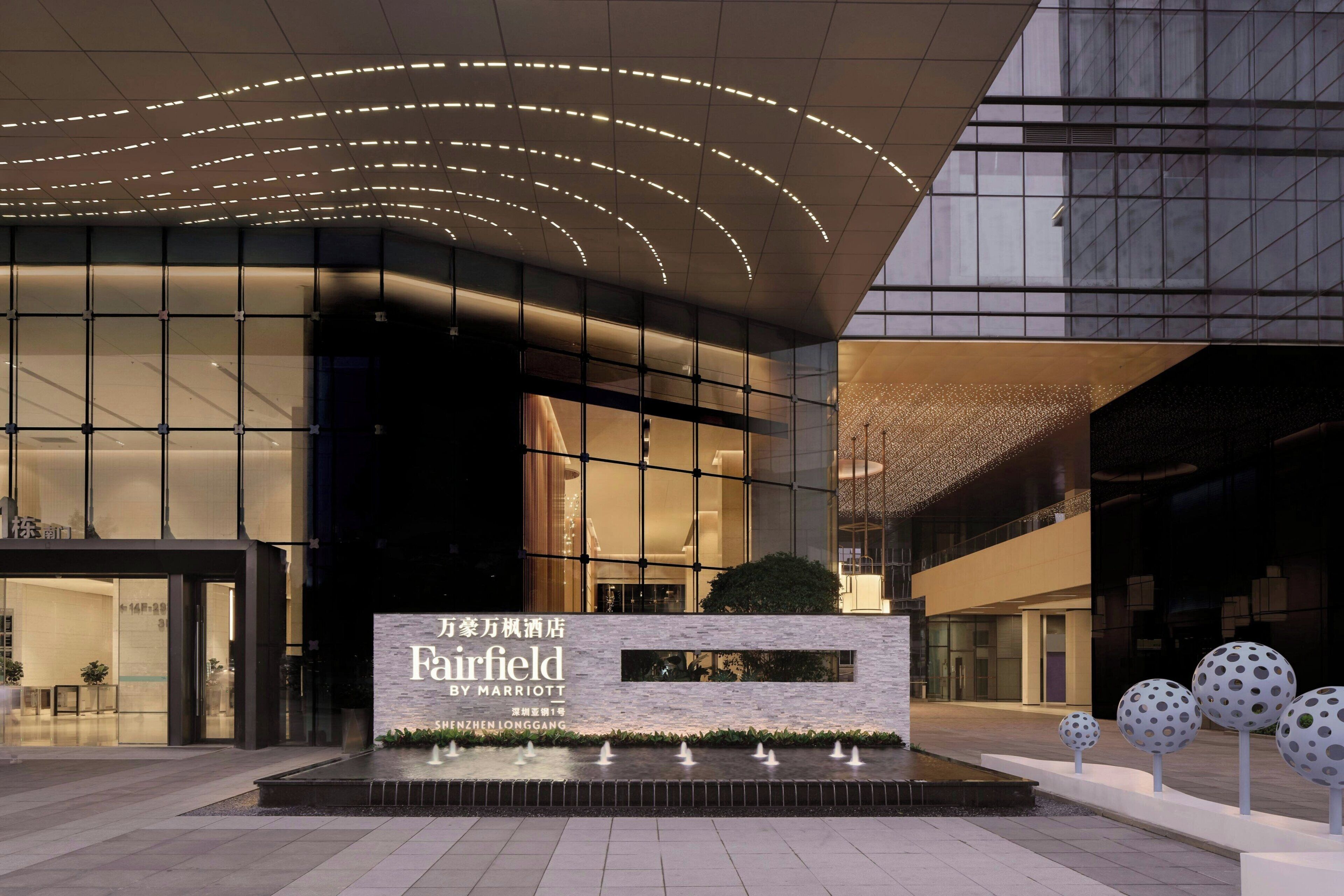 A modern hotel entrance with a large sign, a water feature, and a unique architectural design.