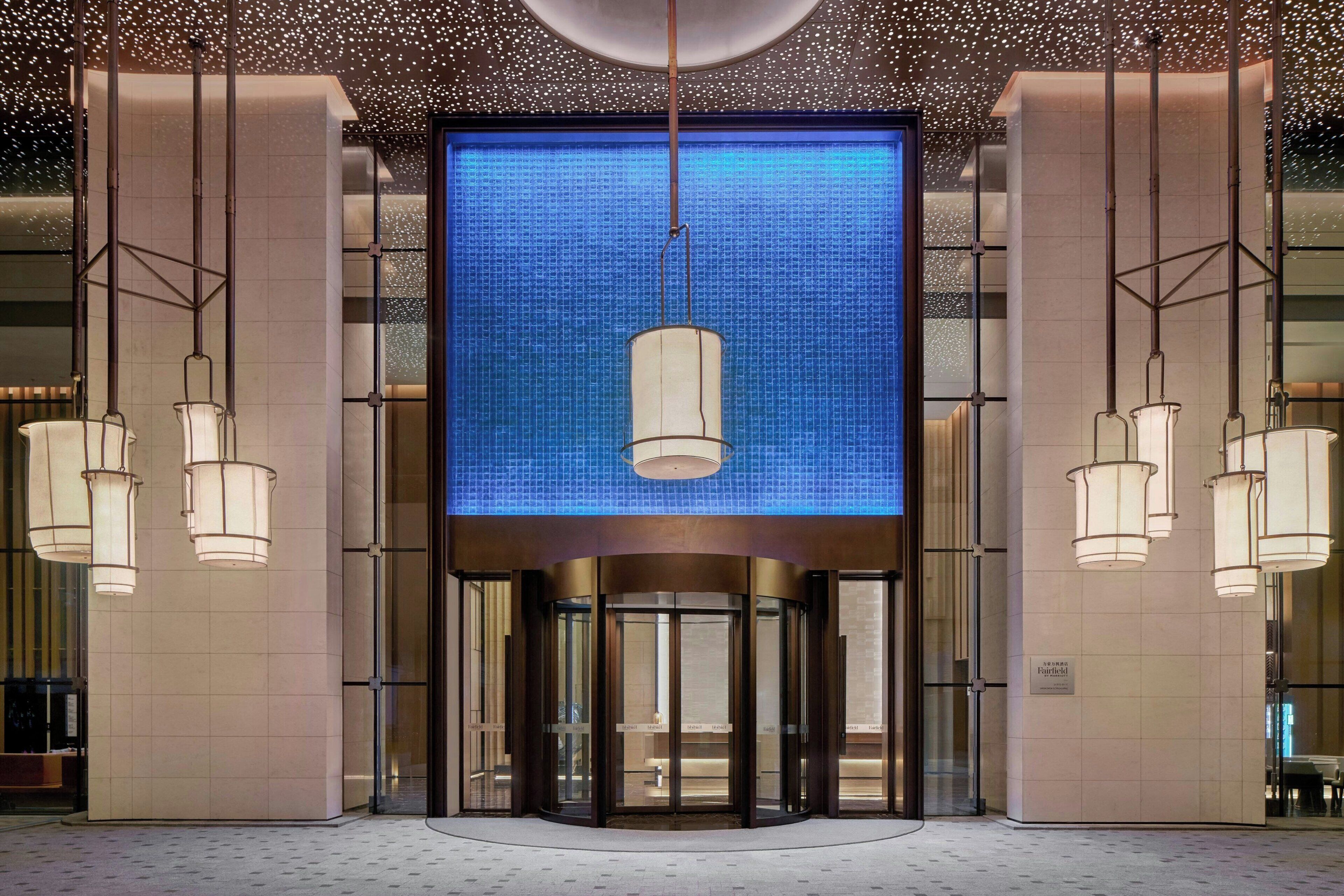 Entrada de hotel con puerta giratoria, una gran pantalla iluminada y lámparas colgantes.