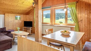 Interior de una cabaña de madera con una mesa de comedor, un sofá, un televisor y un ventanal con vistas a un bosque.