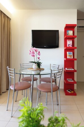Ein Essbereich mit einem Glastisch, Metallstühlen, einer Topforchidee, einem roten Bücherregal und einem an der Wand befestigten Fernseher.