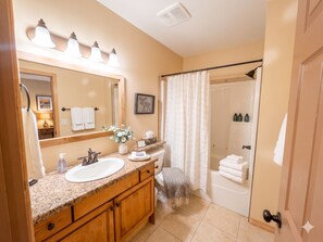 Um banheiro com pia, espelho, bancada, chuveiro e porta-toalhas.