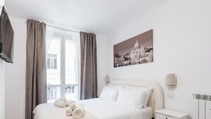A hotel room with a bed, curtains, a wall-mounted TV, a painting, and a radiator.