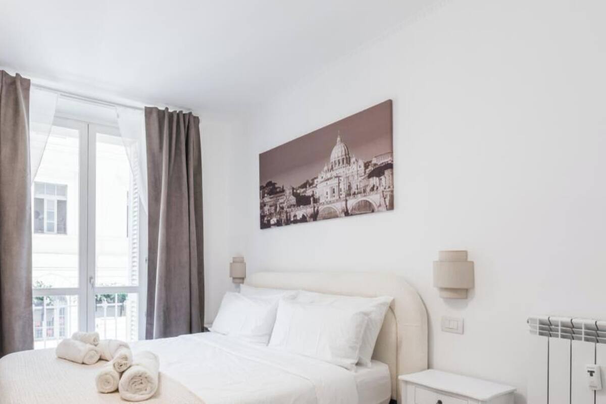 A hotel room with a bed, curtains, a wall-mounted TV, a painting, and a radiator.