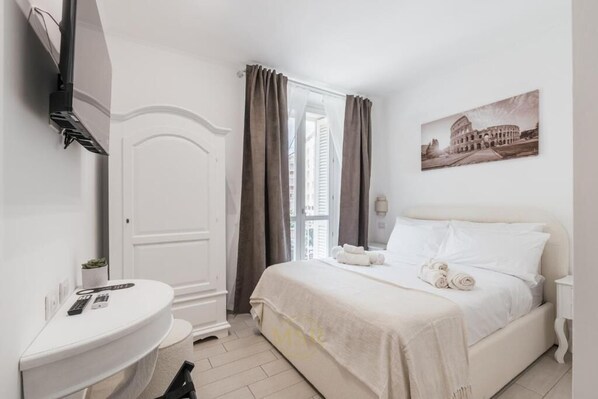 A hotel room with a bed, a desk, a chair, a window with curtains, and a picture of the Colosseum. - My Suite Rome (Rome)