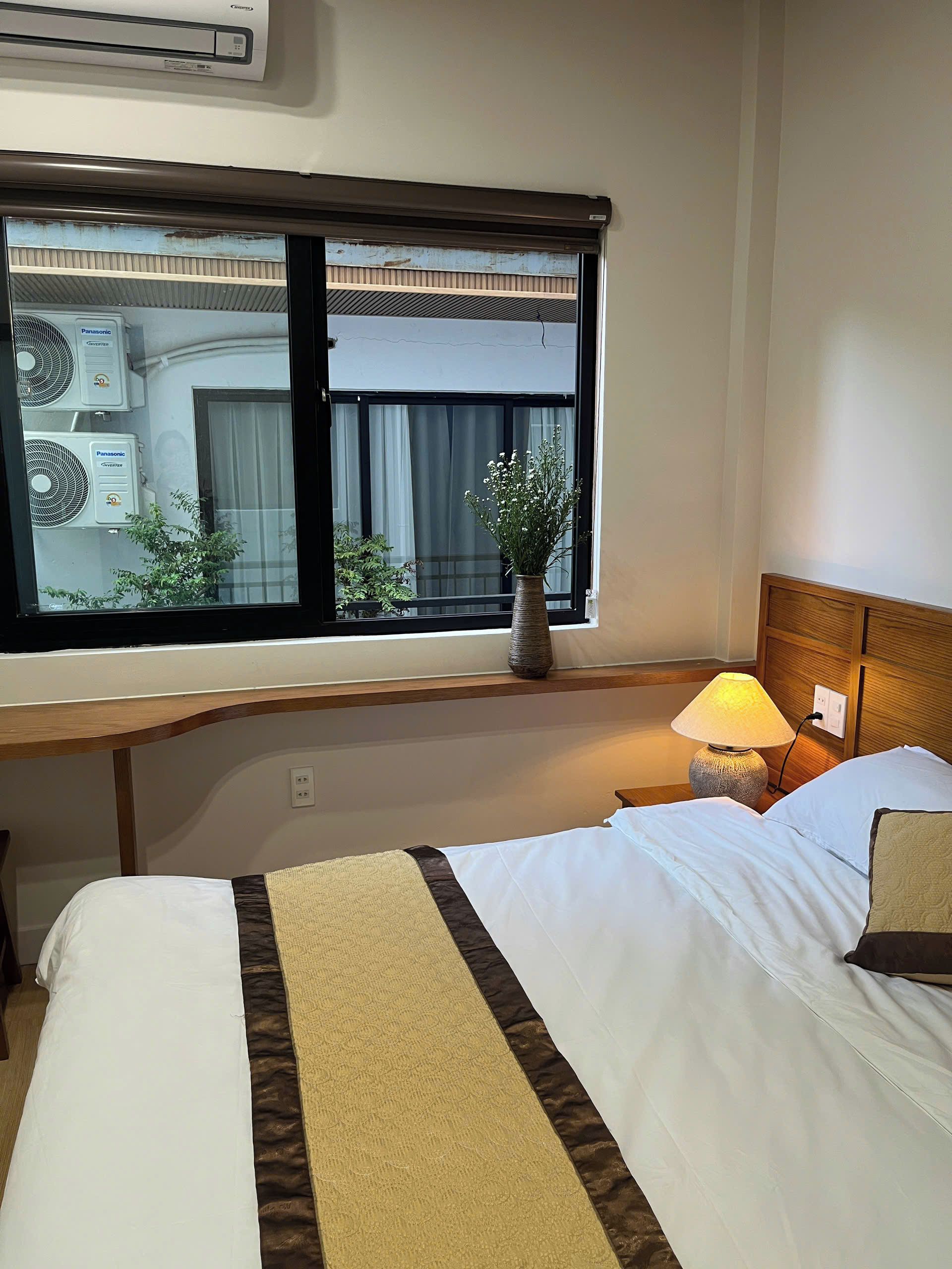 Una habitación de hotel con cama, lámpara de noche, ventana con cortinas e instalación de aire acondicionado.