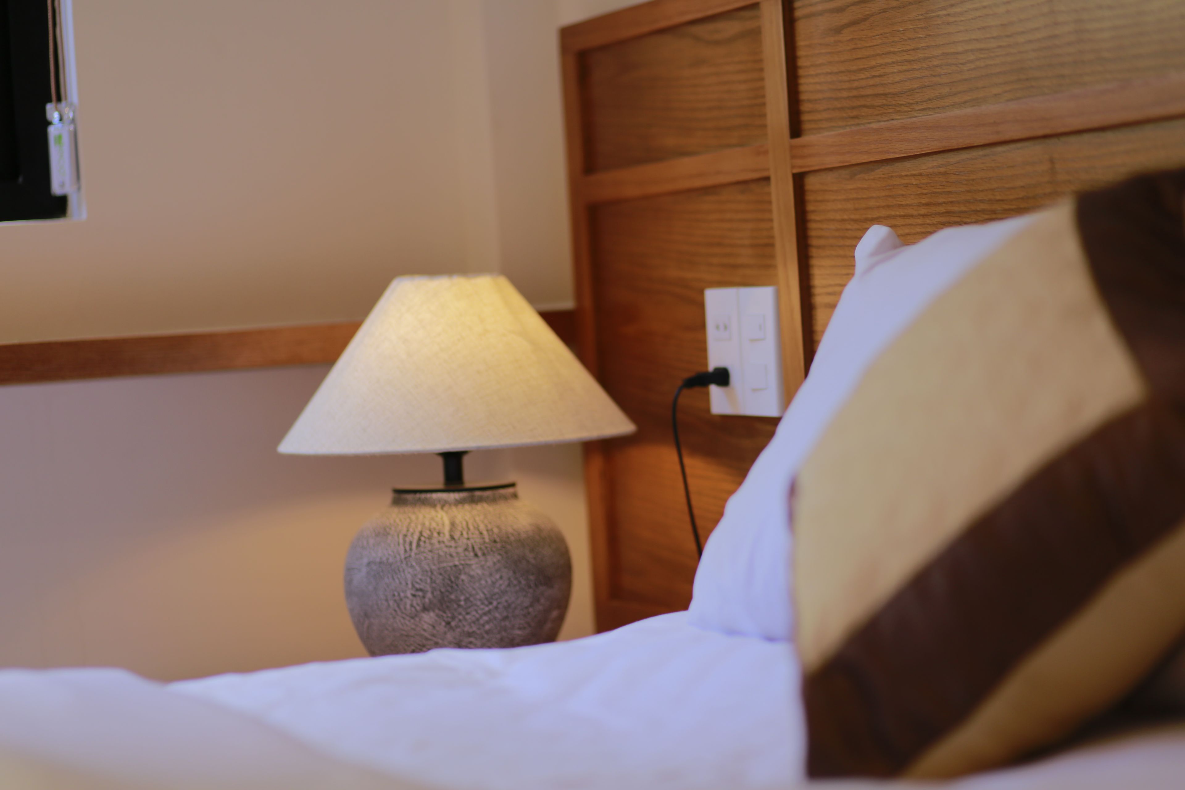 Una cama con ropa de cama blanca y una almohada marrón, una lámpara con base texturizada y un cabecero de madera con toma de corriente.