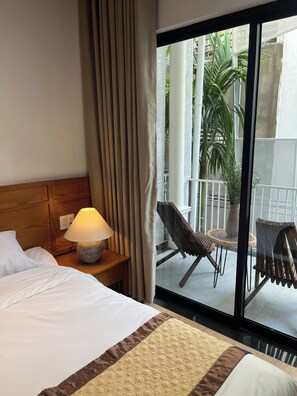 Une chambre d’hôtel avec un lit, une table de chevet avec une lampe, et une vue sur le balcon extérieur, qui est aménagé avec des chaises et un pot de fleurs.