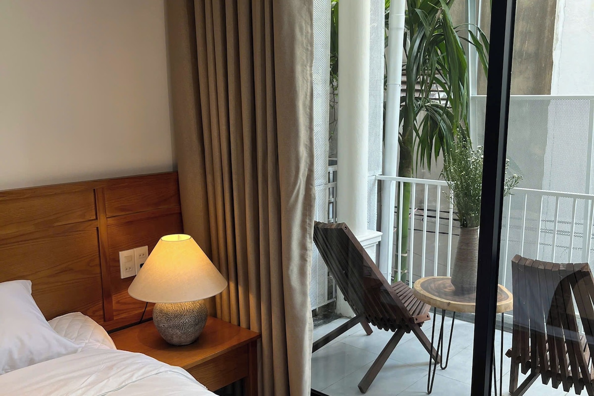 A hotel room with a bed, a nightstand with a lamp, a view of the outdoor balcony with chairs and a potted plant.