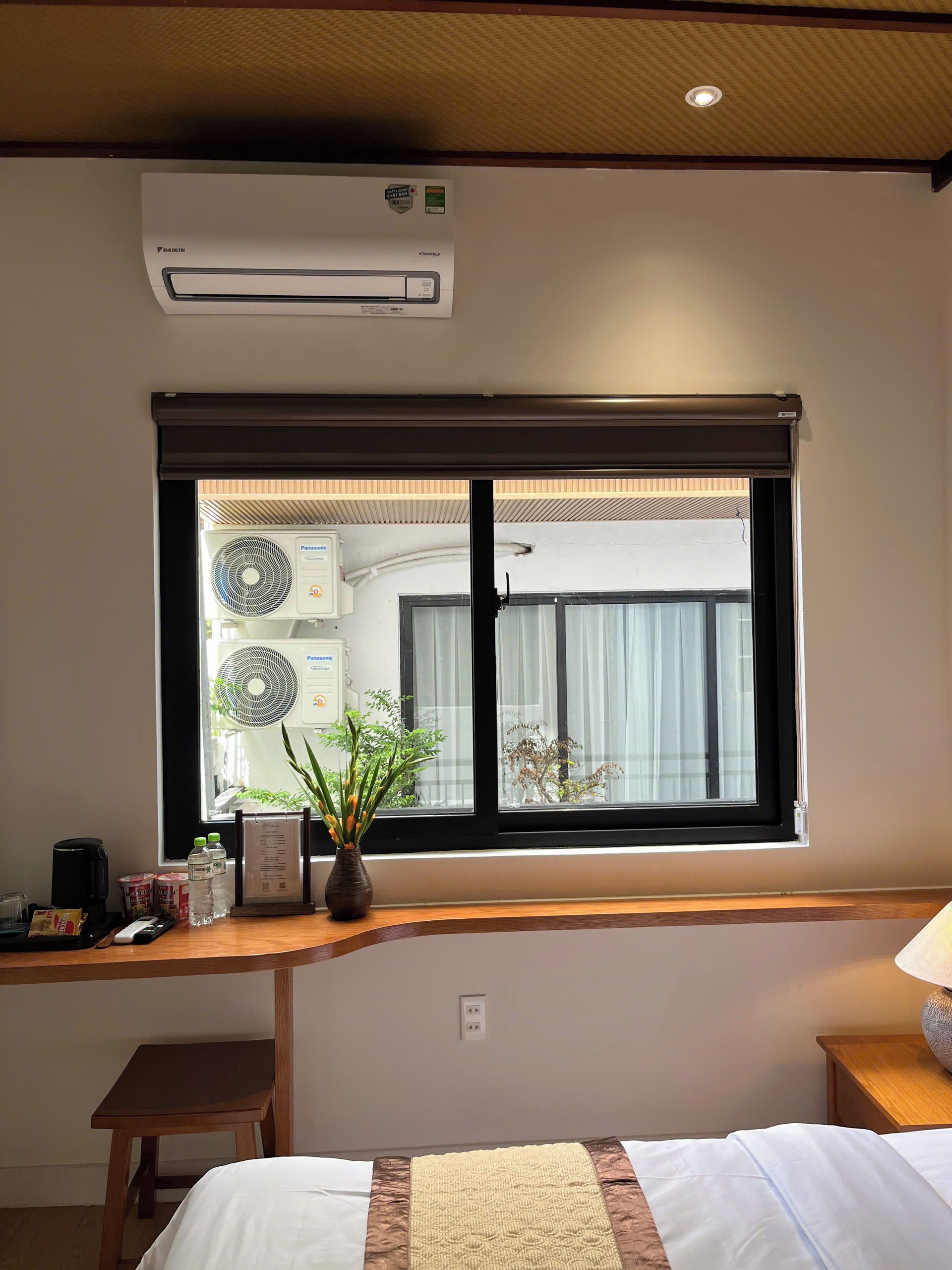 Una habitación de hotel con una cama, un escritorio con una botella de agua, un florero con plantas y una ventana con persianas.