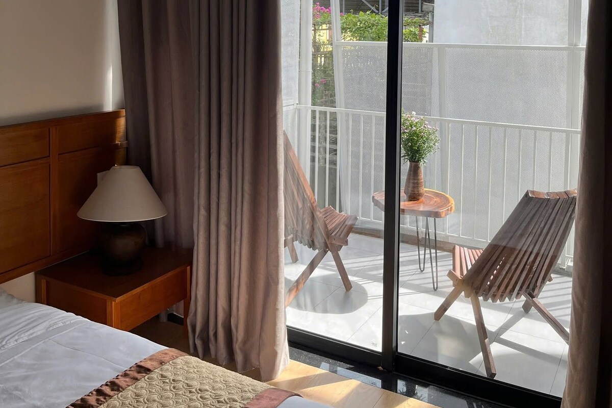 A hotel room with a bed, a nightstand, a lamp, a large sliding glass door leading to an outdoor area with a table and chairs.
