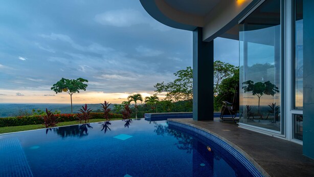 Une piscine à débordement moderne offrant une vue dégagée sur le paysage, un patio entièrement vitré et un jardin bien entretenu.