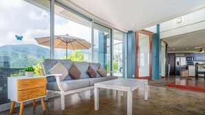 Un salon moderne avec un canapé, une table basse et une table d’appoint, de grandes fenêtres offrant une vue sur les montagnes, et un coin cuisine visible à travers une porte ouverte.