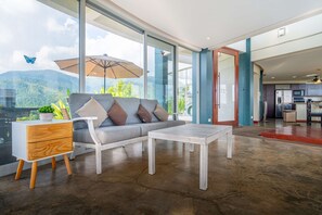 Una sala moderna con sofá, mesa de centro y mesita auxiliar, ventanas amplias con vista a montañas, y un área de cocina visible a través de una puerta abierta.