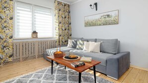 Ein Wohnzimmer mit grauem Sofa, einem Holztisch mit Buch und Korb, einem Fenster mit Jalousien und einer Wandleuchte.