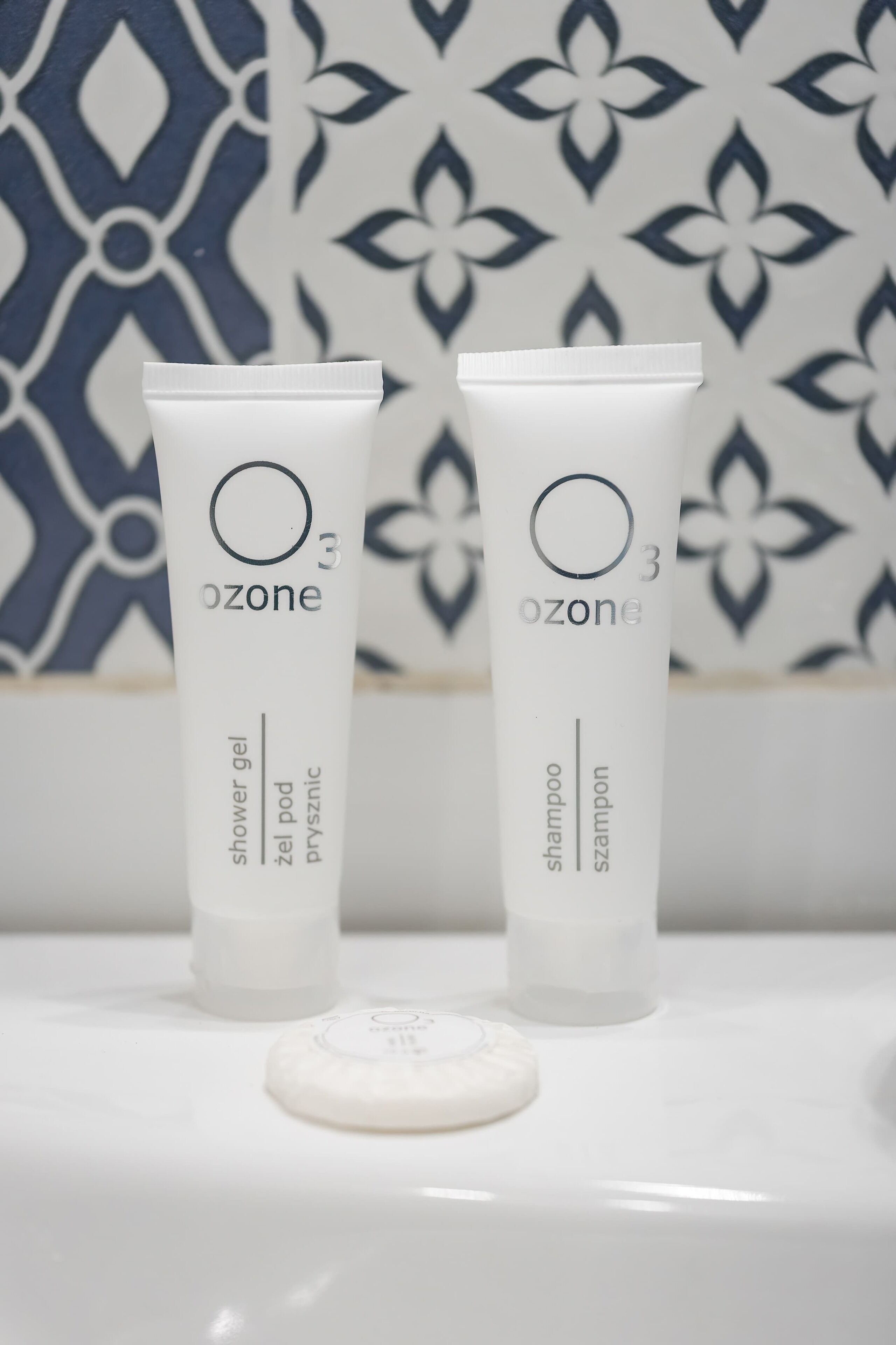 Two white Ozone shower gel tubes and a white soap bar on a white surface with a patterned blue and white background.