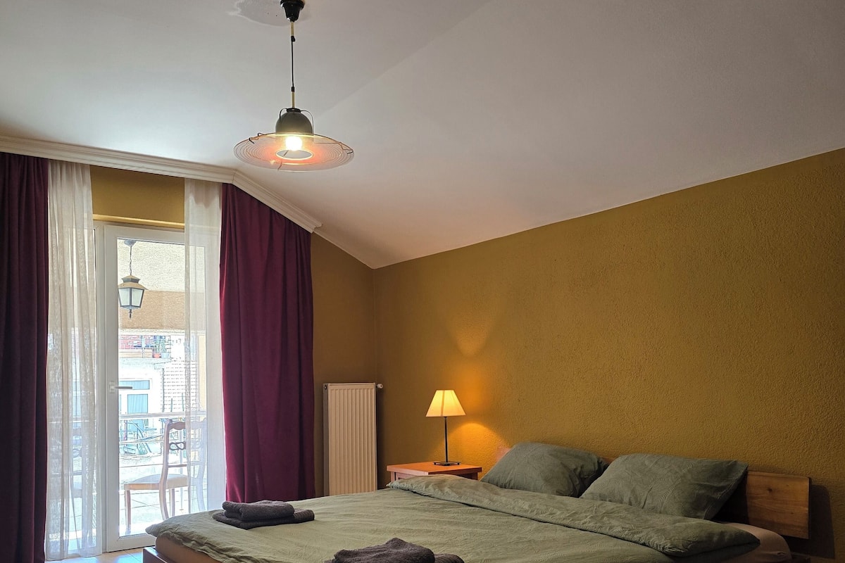 Un dormitorio con cama, mesita de noche, lámpara, ventana con cortinas y vistas al exterior.