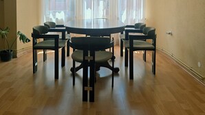 Un coin repas avec une table ronde, six chaises, un luminaire suspendu, de grandes fenêtres avec des rideaux légers et un tableau encadré au mur.