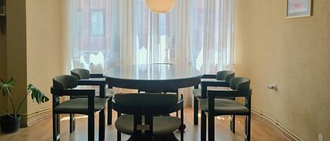 A dining area with a round table, six chairs, a hanging light fixture, large windows with sheer curtains, and a framed picture on the wall.