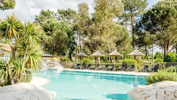 Uima-allas, jossa on aurinkotuoleja ja olkikattoisia aurinkovarjoja, ja jonka ympärillä on puita ja kiviä.