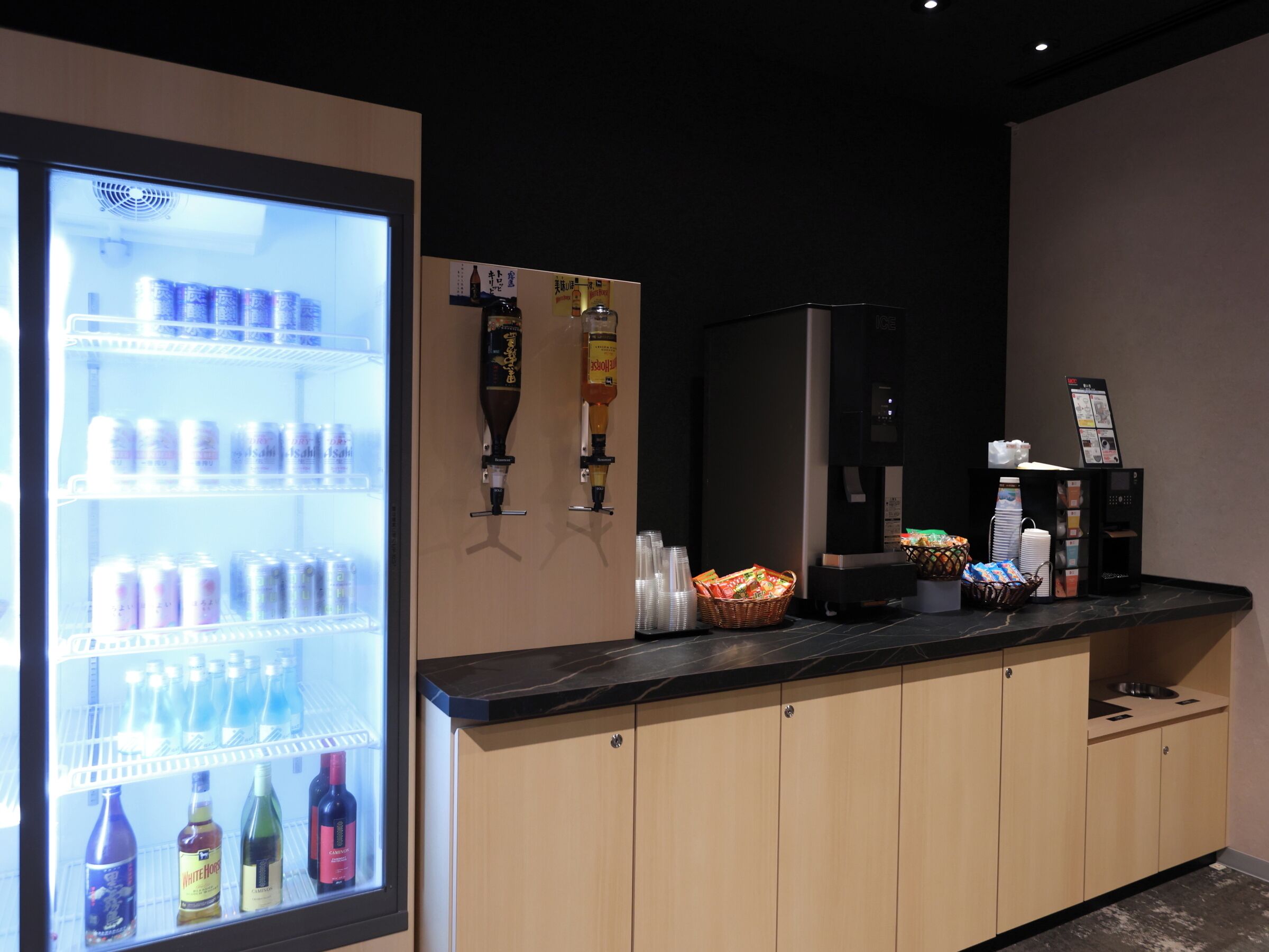 A hotel bar area with a refrigerated beverage display, a coffee machine, and a beverage dispenser.