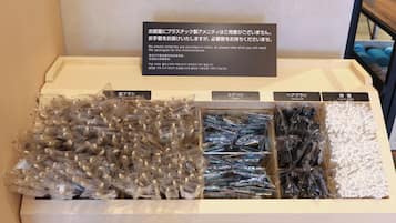 Esposizione di articoli da toilette in un contesto alberghiero, tra cui rasoi, spazzole per capelli e batuffoli di cotone, con un cartello che si scusa per l'assenza di articoli in plastica in camera.