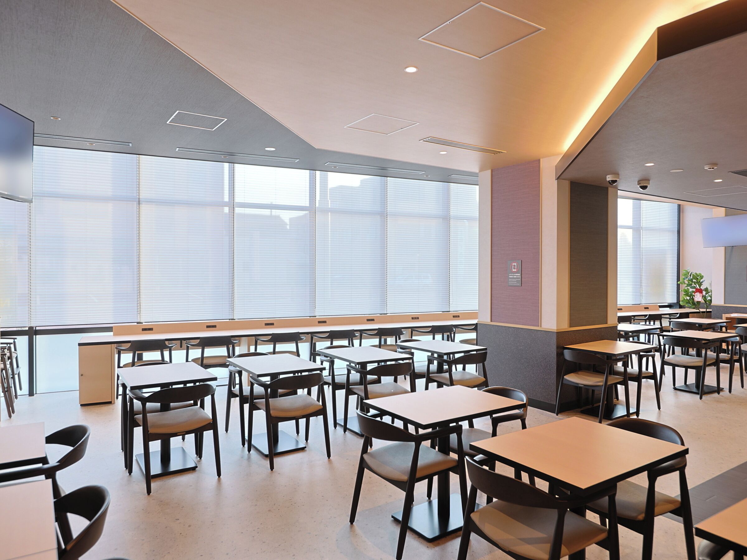 A modern dining area with tables and chairs, large windows, and recessed ceiling lighting.
