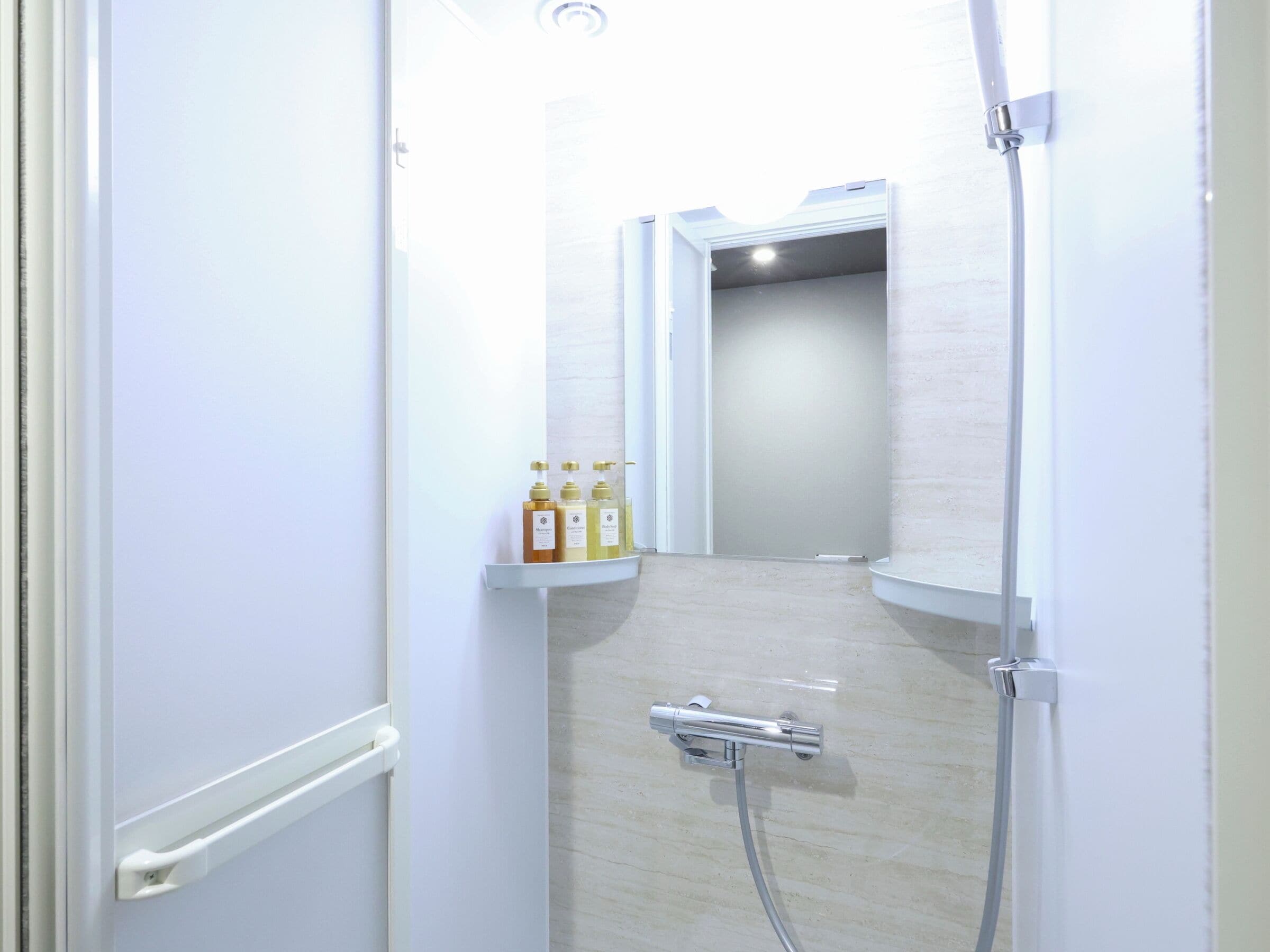 A narrow, white-walled bathroom with a shower, a toilet, and a shelf with toiletries.