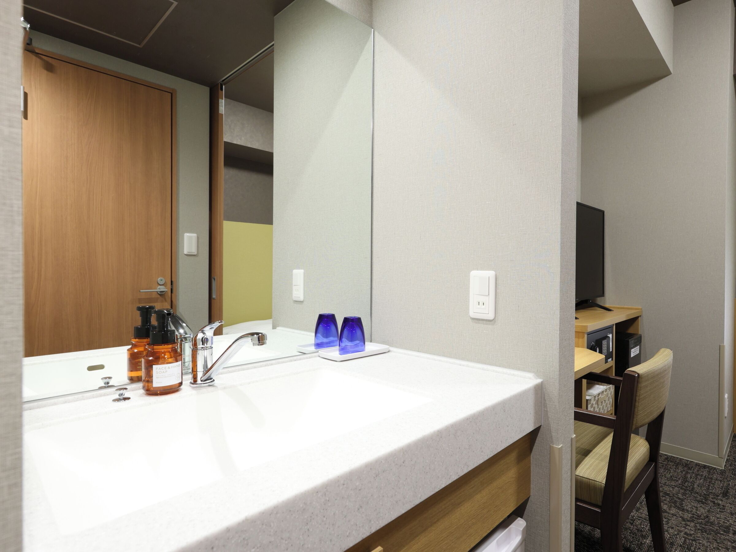 A hotel bathroom with a sink, mirror, toiletries, and a chair.