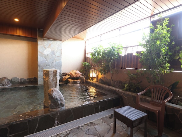 A stone hot tub with a waterfall feature, surrounded by plants and a wooden chair.