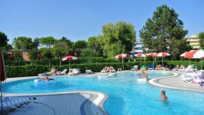 Piscina con sillones de descanso y sombrillas, rodeada de vegetación y edificios.