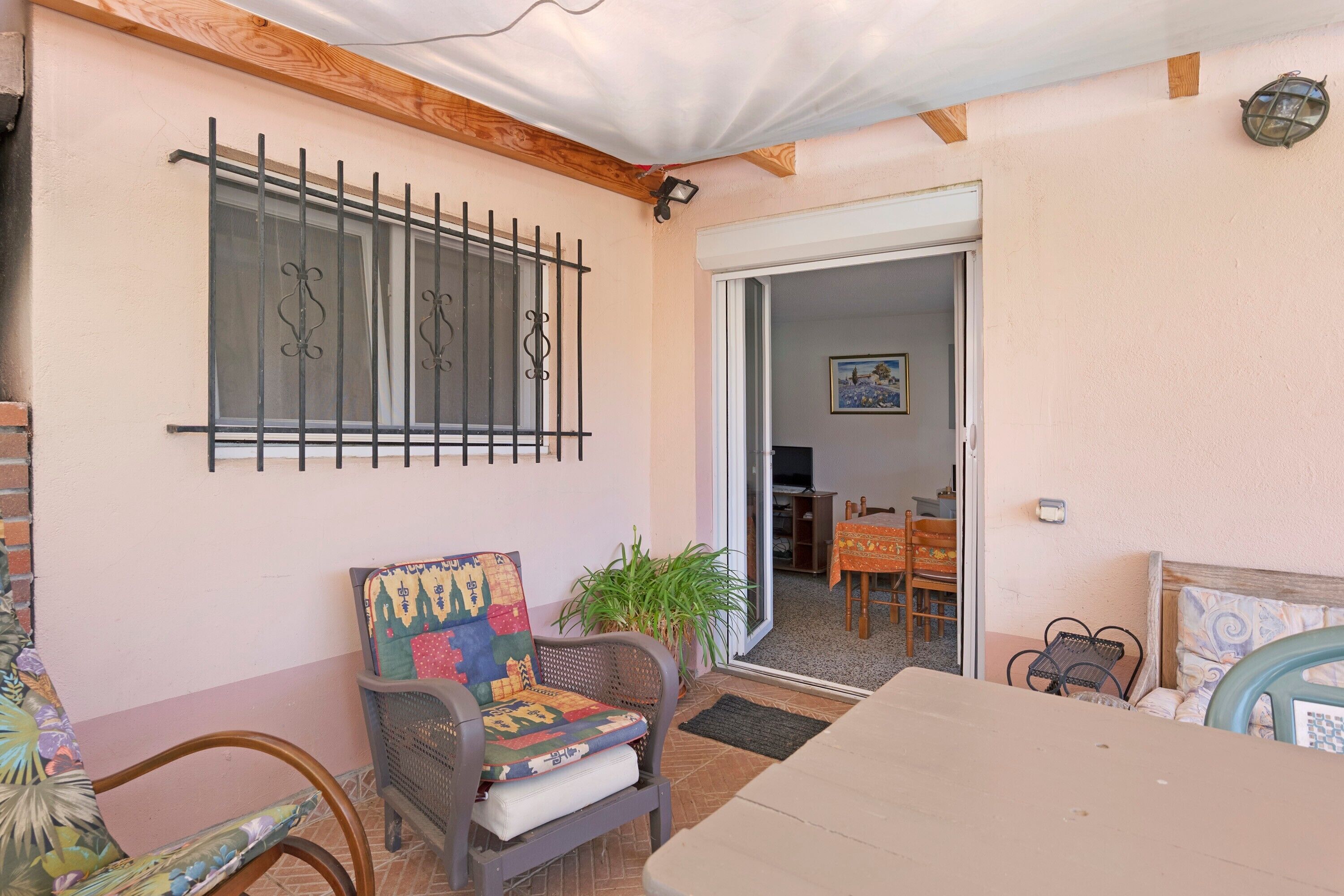 A room with a wooden ceiling, a window with metal bars, a chair with a colorful cushion, a potted plant, and a table.