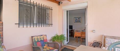 A room with a wooden ceiling, a window with metal bars, a chair with a colorful cushion, a potted plant, and a table.