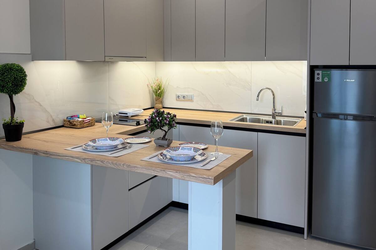A modern kitchen with grey cabinets, a central island with a vase and a potted plant, a stainless steel refrigerator, and a built-in sink.