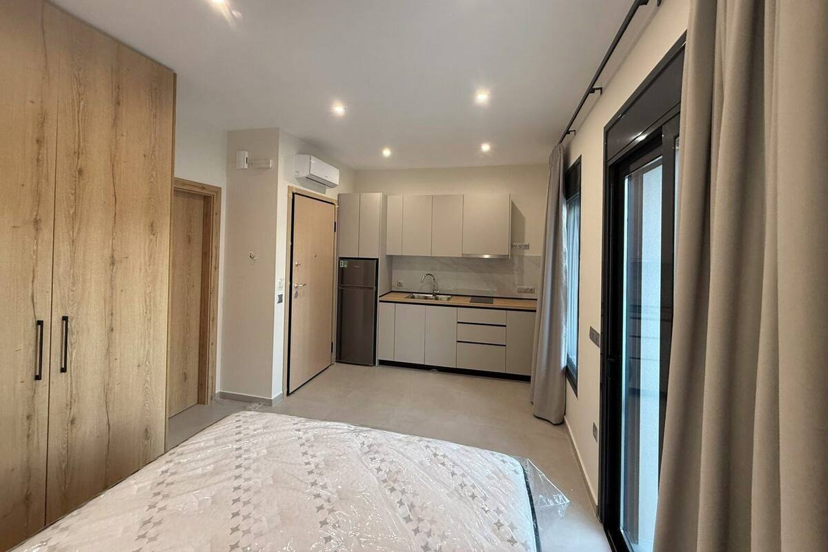 A modern bedroom with a large bed, a wooden wardrobe, a kitchenette, and a sliding glass door to the right.
