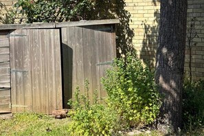 Un abri en bois avec une porte entrouverte, entouré de verdure et bordé d’un mur de briques.